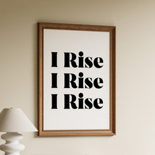 Load image into Gallery viewer, A framed wall art print with the phrase 'I Rise I Rise I Rise' in bold black text on a white background, hanging above a wooden shelf with books and a white lamp in a styled interior setting.