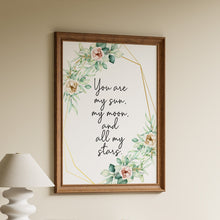Load image into Gallery viewer, A framed wall art print with a floral watercolor border and the handwritten quote 'You are my sun, my moon, and all my stars' hangs on a beige wall above a wooden shelf with books and a white lamp.