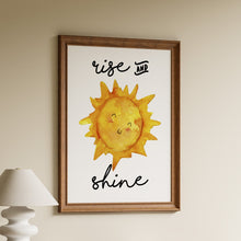 Load image into Gallery viewer, Rise and shine in black hand-lettered script around a cute smiling yellow watercolour sun on a white background in a wooden frame