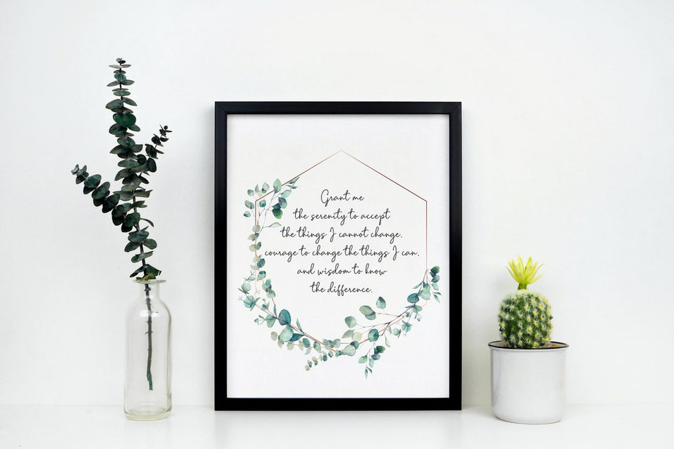 A non-religious serenity prayer print with a watercolor style eucalyptus wreath in a black frame. The framed print is between a small cactus and a glass jar with a sprig of eucalyptus.