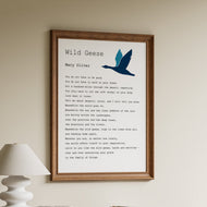 Framed poem titled 'Wild Geese' by Mary Oliver on a wall above a wooden shelf with books and a lamp.