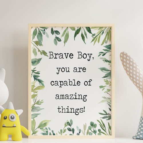 May include: A framed print with a white background and green watercolor leaves around the edges. The text on the print reads 'Brave Boy, you are capable of amazing things!' The print is sitting on a white shelf with a white stuffed cat, a yellow stuffed monster, and a blue and white stuffed bunny on the shelf. The text 'Frame Not Included' is printed below the print.