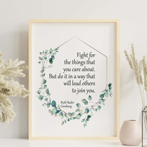 May include: A framed print with a white background and a green leafy border. The print features a quote by Ruth Bader Ginsburg that reads 'Fight for the things that you care about. But do it in a way that will lead others to join you.' The print is displayed on a white shelf with decorative items.