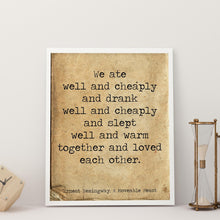 Load image into Gallery viewer, May include: A white framed print with a vintage, brown background. The print features a typewriter font quote that reads: &#39;We ate well and cheaply and drank well and cheaply and slept well and warm together and loved each other.&#39; The quote is attributed to Ernest Hemingway and the book &#39;A Moveable Feast.&#39; The print is sitting on a white surface with two gold hourglass sand timers.
