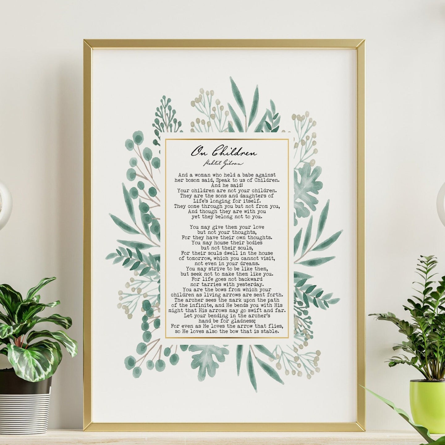 May include: A framed print with a gold frame and a white background. The print features a poem titled 'On Children' by poet John Greenleaf Whittier. The poem is surrounded by a watercolor illustration of green leaves and flowers. The print is displayed on a wooden shelf with two green plants and two light bulbs hanging from the ceiling.